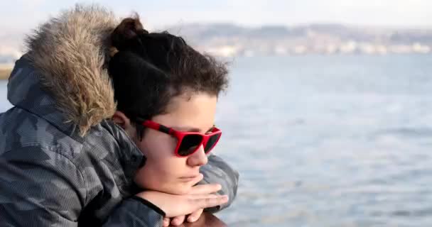 一个戴着太阳镜 穿着冬衣 独自坐在海边的英俊忧伤少年的画像 — 图库视频影像