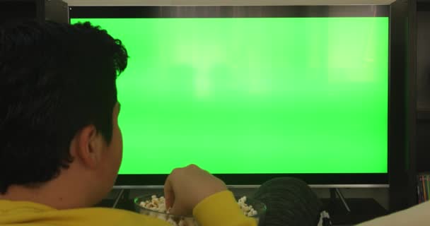 坐在沙发上看电视绿屏和在家里吃零食的十几岁男孩的倒影 — 图库视频影像
