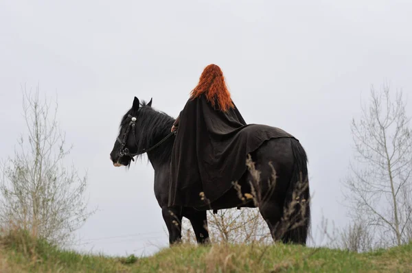 Tapfere Frau mit roten Haaren im schwarzen Mantel auf einem Friesenpferd — Stockfoto