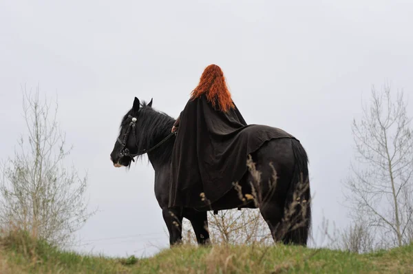 Friesian 말에 검은 망 토에 붉은 머리를 가진 멋진 여자 — 스톡 사진