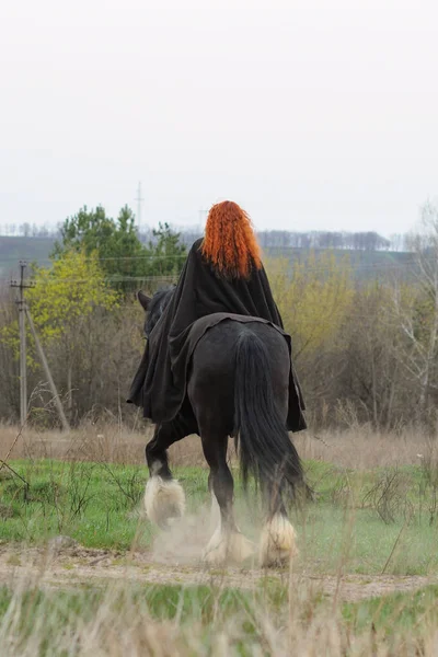 Dzielna kobieta z rude włosy w czarny płaszcz na Koń fryzyjski Obrazy Stockowe bez tantiem