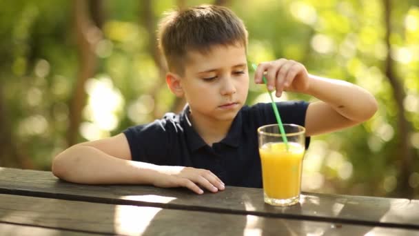 Chłopiec pije sok pomarańczowy przy stole w przyrodzie. Młody chłopak pije sok pomarańczowy na zewnątrz. — Wideo stockowe
