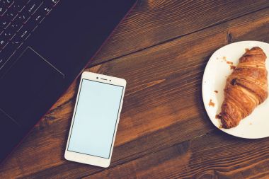 Dizüstü bilgisayar, akıllı telefon ve kruvasan ahşap masa üzerinde çalışma alanı