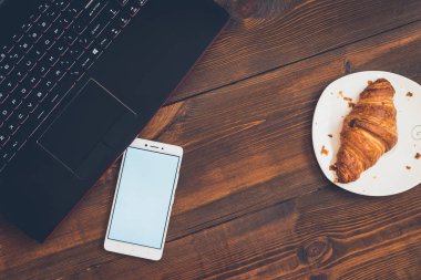 Dizüstü bilgisayar, akıllı telefon ve kruvasan ahşap masa üzerinde çalışma alanı