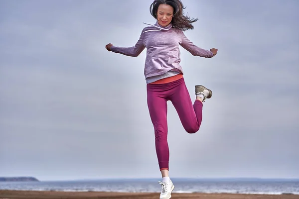 Young woman with black hair jumps while jogging. A woman is engaged in gymnastics in the spring morning on the sandy bank of a large river. Cloudy morning.
