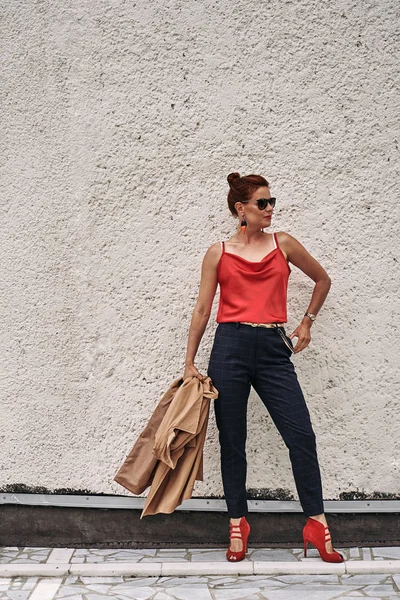 Portrait of a red-haired middle-aged woman in a red blouse  and in sunglasses  standing against the wall. Cloudy summer day. Close-up.