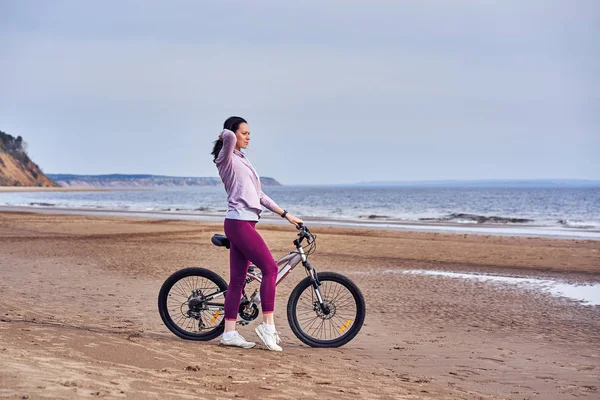 Young slim brunette woman on a bike ride on the sandy shore of a large river. Cloudy summer day.