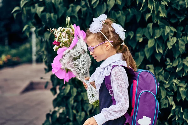 A girl child in school uniform with bows, glasses and a school bag on his back goes to the first class of the school on September 1st. The girl is holding a bouquet of flowers. Close-up.
