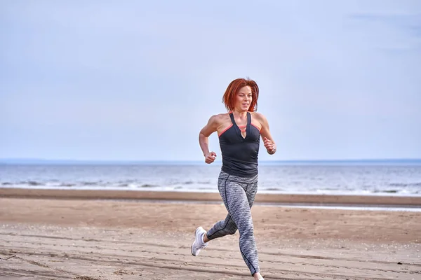 A red-haired middle-aged woman in sportswear runs along the sandy shore of a large river on a cloudy summer morning. Healthy lifestyle.