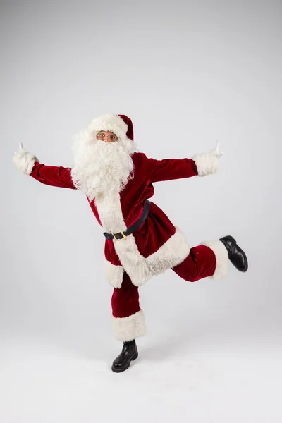 Santa Claus en gafas y traje rojo equilibrado en una pierna sobre fondo blanco y mirando a la cámara . — Foto de Stock