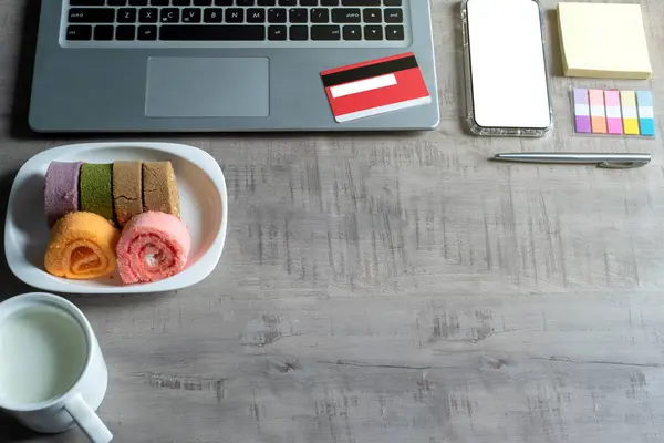 Dizüstü bilgisayar, akıllı telefon, kredi kartı, bir fincan süt, reçel rulo, kağıt not, kalem, ahşap masa üzerinde iş, ticaret, finans, eğitim konsepti ve tasarım