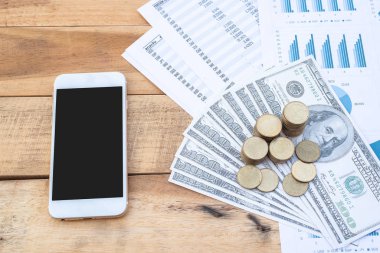 Flat Lay of stack of coins on dollar banknote, smartphone, blue pastel chart paper on the wooden table. Business, finance, marketing, e-commerce concept and design.