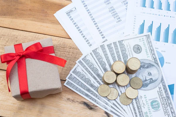 Flat Lay of stack of coins on dollar banknote, red ribbon giftbox, blue pastel chart paper on the wooden table. Business, finance, marketing, e-commerce concept and design.