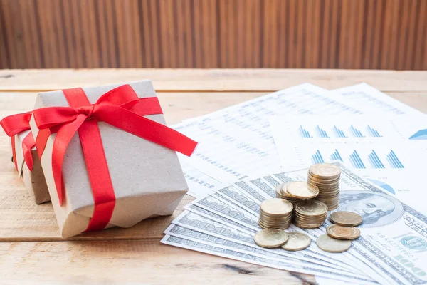 Flat Lay of stack of coins on dollar banknote, red ribbon giftbox, blue pastel chart paper on the wooden table. Business, finance, marketing, e-commerce concept and design.