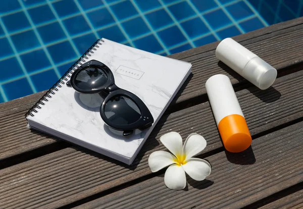 Sunscreen, planner, sunglasses and tropical flower near swimming pool