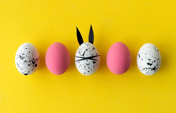 Composición de Pascua con huevos de colores y huevo con orejas de conejo y cara sobre fondo amarillo — Foto de Stock