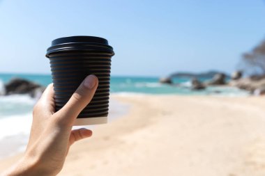 Kartondan kahve fincanı taşıyan kadın eli, deniz kenarında. Rahatla. Tatil kavramı..