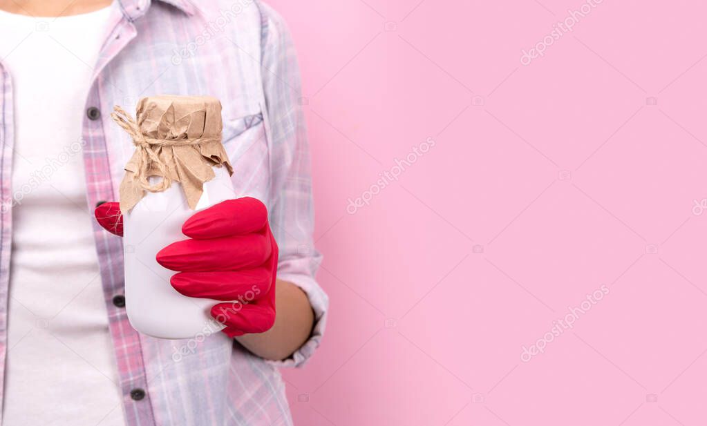 Womans hand in a rubber glove holding white bottle on pink background. Stay safe home delivery concept.