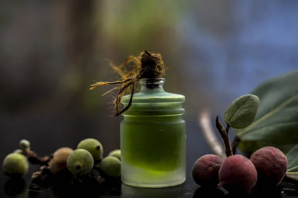 Close up shot of essential banyan tree oil in a transparent glass bottle along with some banyan fruit and its leaf on a black glossy wooden surface. Horizontal shot.