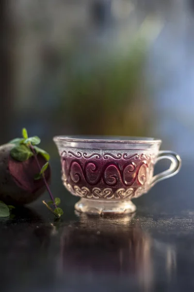 Best detoxify drink on a black glossy surface in a glass cup. Beetroot tea in a transparent glass cup on a black surface with a raw beet and some mint leaves. Vertical shot with blurred background.