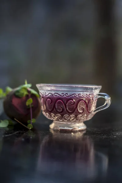 Best detoxify drink on a black glossy surface in a glass cup. Beetroot tea in a transparent glass cup on a black surface with a raw beet and some mint leaves. Vertical shot with blurred background.