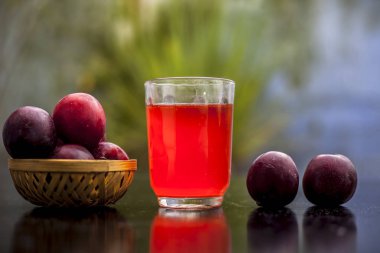 Siyah parlak yüzeyde kahverengi renkli bir sepetin içinde ham organik kırmızı erikler seçici odaklı ve bulanık arkaplanlı şeffaf bir bardakta taze meyve suyu çıkartılıyor..