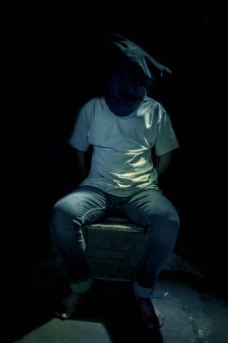 Adult human sitting on a rusted metal chair wearing a black colored bag covering his face.In dark gothic colors.
