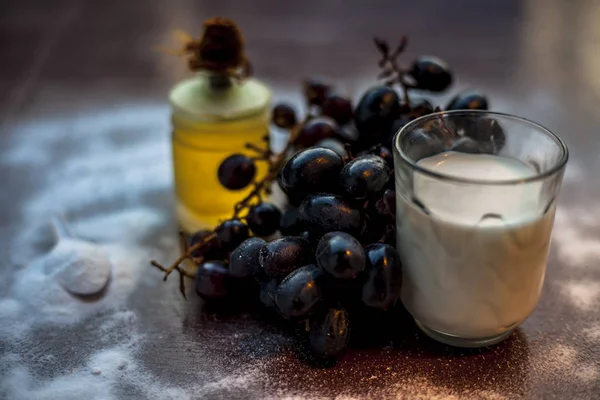 Natural fruit face mask cleanser on the brown colored surface consisting of a bunch of grapes, baking soda, olive oil, and some milk. Shot of grapes spread baking soda olive oil and milk in a glass.