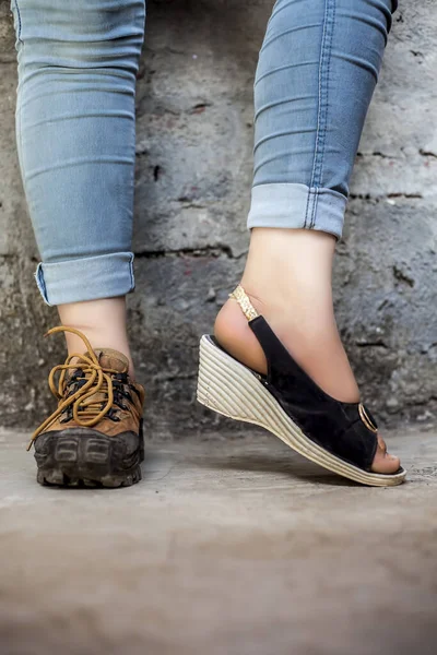 Female foot wearing hiking boots in one foot and sandals in another and posing against a brick wall wearing jeans.