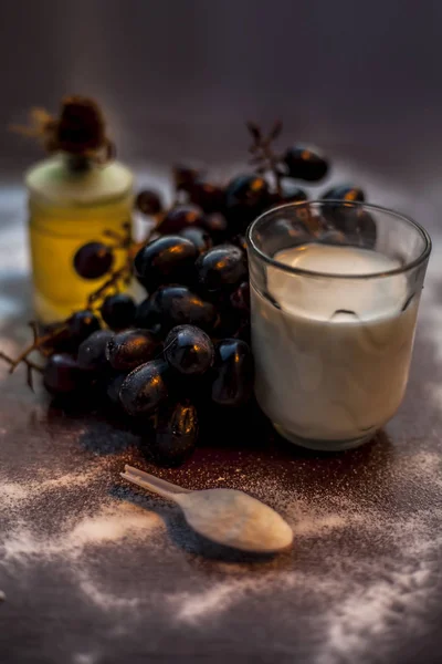 Natural fruit face mask cleanser on the brown colored surface consisting of a bunch of grapes, baking soda, olive oil, and some milk. Shot of grapes spread baking soda olive oil and milk in a glass.