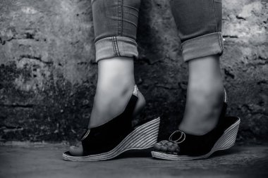 Mavi kot pantolon ve dikiz ayağında sandalet giyen zarif genç bir kızın siyah beyaz arka planda tuğla duvara karşı bacak bacak bacak üstüne atarak poz vermesi..