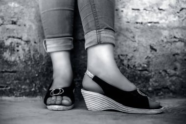 Mavi kot pantolon ve dikiz ayağında sandalet giyen zarif genç bir kızın siyah beyaz arka planda tuğla duvara karşı bacak bacak bacak üstüne atarak poz vermesi..