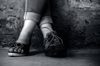 Bir ayağında dikiz parmaklı sandaletler, diğerinde yürüyüş botları olan mavi kot pantolon giyen genç bir kızın zarif ayağı. Siyah beyaz renkli yatay çekim..