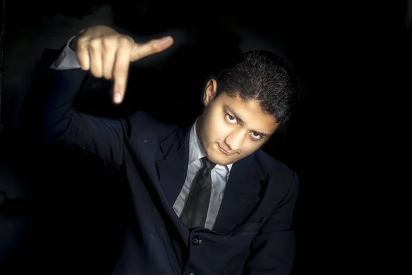 Portrait shot of an arrogant businessman posing on a black colored background in a blue suit with one hand pointing downwards showing arrogance on his face. Horizontal shot.