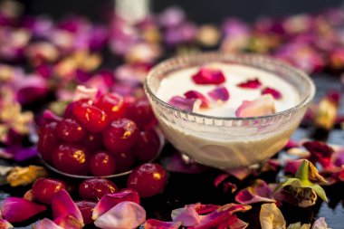 Siyah ahşap bir tahtanın üzerinde, biraz kiraz ve yoğurt içeren cilt parlatıcı yüz maskesi. Yüzeydeki tüm malzemelerle birlikte birkaç gül yaprağı. Yatay çekim.
