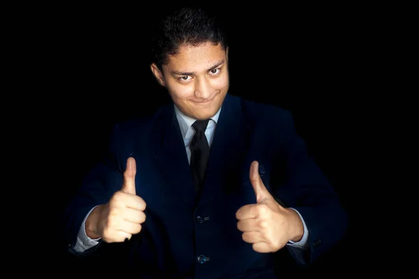 Portrait shot of smiling charming young businessman in dark blue shirt and Wishing Best Wishing Good luck with thumbs up Or Best of luck hand gesture over a black colored background. Horizontal shot.