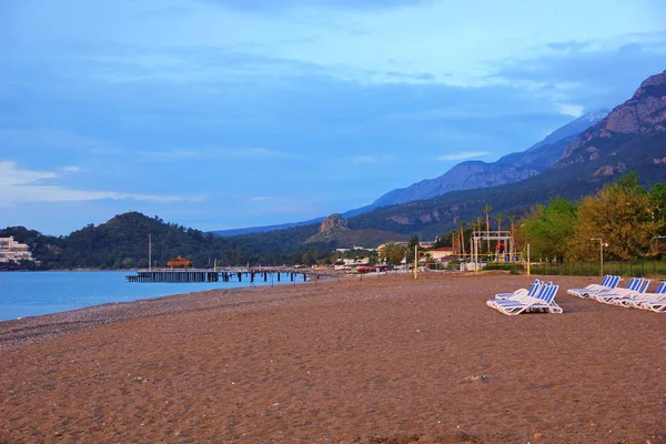 Sabahın erken saatlerinde ayakta sandalye, bir dizi Türk resort kum çakıl plaj dağlar çevreleyen resort, palmiye ağaçları, deniz kenarına, birkaç bulutlu gökyüzü