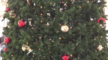 Oyuncaklarla süslenmiş geniş bir Noel ağacı ve yanıp sönen bir çelenk, kırmızı, altın toplar, kenarları görünür insanlar, Noel telaş içinde