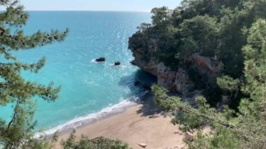 Kumlu sahil ve deniz berrak turkuaz suyla, dalgalar kumsalda akıyor, yeşil bitki örtüsü, çam ağaçları, denizin sesi.