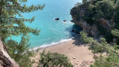 Kumlu sahil ve deniz berrak turkuaz suyla, dalgalar kumsalda akıyor, yeşil bitki örtüsü, çam ağaçları, denizin sesi.