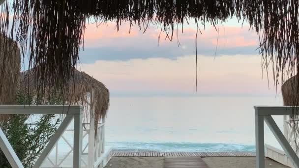 海の茅葺き屋根のビーチテントからの眺め 柔らかい繊細な色 岩のビーチ ピンクと白の空 — ストック動画