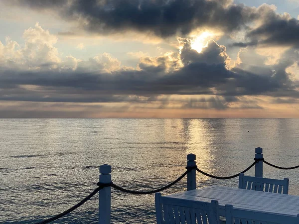 view of the morning sunrise by the sea, beautiful sky with clouds, sun rays break through, partially visible cafe, railing and table with chairs