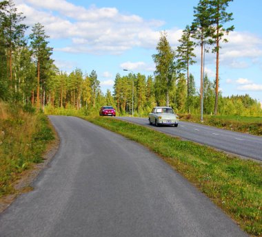 Finlandiya, Imatra, 18 Ağustos 2019. Çam ormanının ortasında bir yol, mükemmel durumdaki eski arabalardan biri, ikinci modern, bisikletçiler için geniş bir şerit, güneşli bir gün.