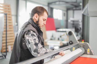 İşçi matbaa teknisyeni, dijital matbaada yarı otomatik yapıştırma makinesiyle köpük panoya büyük kağıt yapıştırıcı yapıştırıyor.