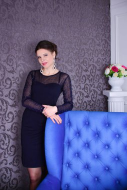 Siyah elbiseli güzel esmer, mavi kanepenin yanında duruyor.