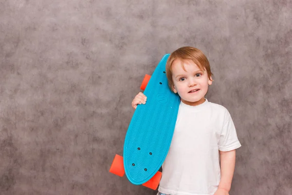 Маленький мальчик в повседневной одежде, держит скейтборд на плече, улыбается, стоя на сером фоне. Концепция продажи спортивного оборудования — стоковое фото