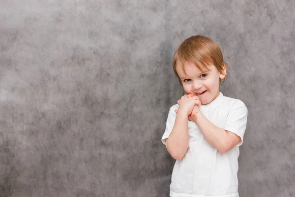 Мальчик со счастливой улыбкой на лице радостно хлопает в ладоши и стесняется. Портрет на сером фоне. Летний стиль — стоковое фото