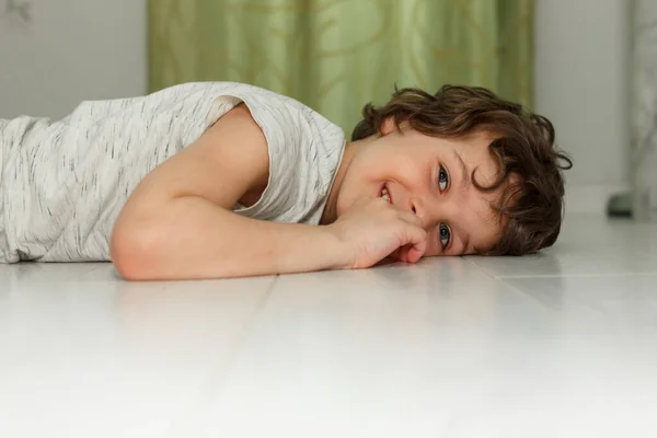 Кудрявый мальчик лежит дома на белом полу и улыбается. Концепция подогрева полов, качественный ламинат — стоковое фото
