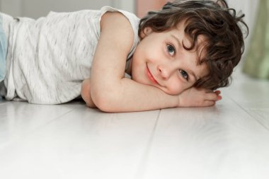 Kıvırcık çocuk evde beyaz zeminde yatıyor ve gülümsüyor. Zemin altı ısıtma ve kaliteli kaplama konsepti..