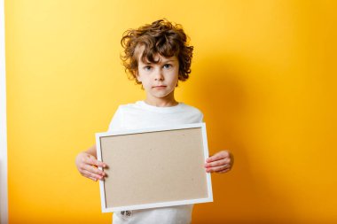 Beyaz tişörtlü kıvırcık çocuk mesaj için boş bir çerçeve tutuyor ve parlak sarı bir arka plan üzerinde parmağını gösteriyor. Beyaz pano ya da afiş - metni doldurmak için boşluk konseptini kopyala. İleti tahtası.
